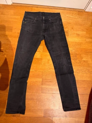 Svarta jeans från Tiger of Sweden - Säljer ett par svarta jeans från Tiger of Sweden med klassisk femficksmodell och raka ben. Jeansen har slitna detaljer och en diskret logga på myntfickan. Materialet är mjukt denim i bomull som ger en avslappnad look. DEFEKT lagning vid skrevet