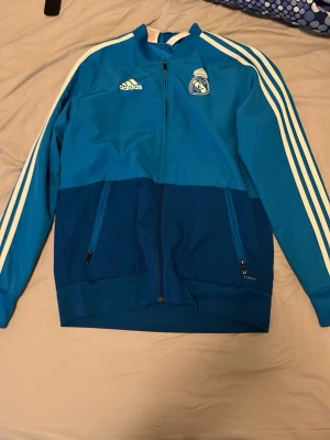 Blå Real Madrid träningsjacka Adidas - Snygg blå träningsjacka från Adidas med Real Madrid-logga på bröstet. Jackan har vita ränder längs ärmarna, dragkedja framtill och två fickor med dragkedja. Tillverkad i ett funktionsmaterial som andas, perfekt för träning eller sport.