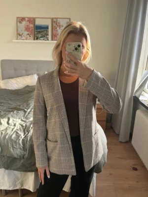 Beige rutig kavaj med klassisk passform - Snygg beige kavaj med rutigt mönster och klassisk krage. Kavajen har två fickor framtill och stängs med dold knapp. Perfekt för en stilren look och passar till både jeans och kostymbyxor.