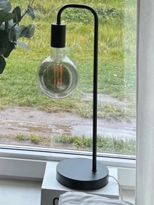 Svart bordslampa med glödlampa - Modern bordslampa i svart metall med rund fot och böjd arm. Lampan har en stor, rund glödlampa med synlig glödtråd som ger ett industriellt intryck. Passar perfekt på fönsterbrädan eller skrivbordet för en stilren look.
