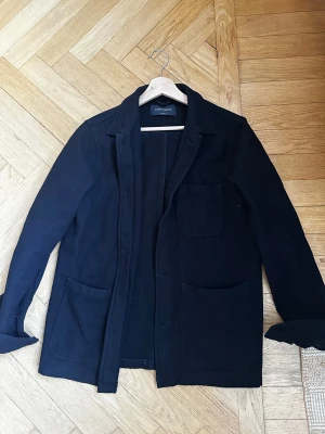 Mörkblå overshirt från A Day's March i ull. - Stilren svart overshirt från A Day's March i storlek medium i ull. Jackan har tre stora utanpåfickor, klassisk krage och rak passform. Materialet är kraftigt och ger en clean look, perfekt för lager-på-lager-stil.