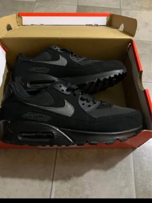 Nike Air Max 90 svarta sneakers - Snygga Nike Air Max 90 sneakers i helsvart med grå detaljer och klassisk siluett. Skorna har en ikonisk synlig Air-enhet i sulan, mesh och syntetmaterial på ovandelen samt Nike-logga på sidan och plösen. Perfekta för dig som gillar streetwear och sportig stil.