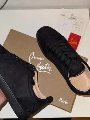 Svarta sneakers från Christian Louboutin - Stilrena svarta sneakers från Christian Louboutin i mocka med klassisk röd sula och diskret logga på innersulan. Skorna har rund tå, snörning och en platt sula. Perfekta för dig som vill ha en lyxig och tidlös sneaker med exklusiv känsla.