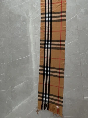 Beige rutig halsduk från Burberry - Klassisk beige halsduk från Burberry med det ikoniska rutmönstret i svart, vitt och rött. Halsduken har fransar i ändarna och är tillverkad i mjukt material som känns lyxigt. Perfekt accessoar för att lyfta din outfit med en tidlös touch.