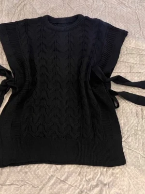 Svart kabelstickad väst med knytband - Snygg svart kabelstickad väst med breda axlar och knytband i sidorna. Perfekt att ha över en långärmad tröja för en cool lager-på-lager-look. Mjukt stickat material och rund halsringning ger en trendig och bekväm stil.