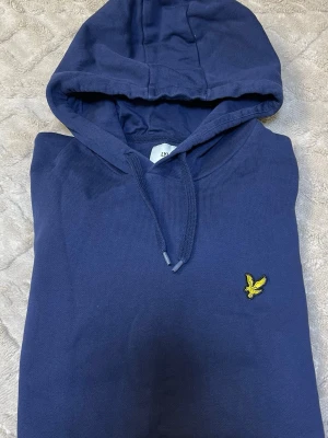 Marinblå hoodie från Lyle & Scott - Snygg marinblå hoodie från Lyle & Scott med klassisk gul logga på bröstet. Hoodien har dragsko i huvan och är tillverkad i mjukt bomullsmaterial. Perfekt för en avslappnad och stilren look.