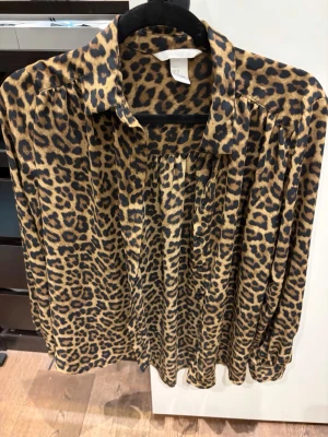 Leopardmönstrad blus från H&M - Snygg leopardmönstrad blus från H&M i mjukt, glansigt material. Blusen har krage, knäppning framtill och långa ärmar med lätt puff vid axlarna. Perfekt för dig som vill sticka ut med ett djurmönster!