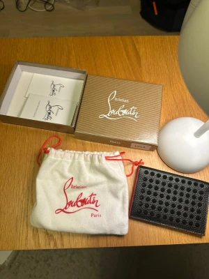 Christian Louboutin plånbok i läder - Snygg plånbok från Christian Louboutin i svart och rött läder. Utsidan har svarta nitar och insidan är klarröd med flera kortfack och präglad logga. Kommer med originalask och dustbag. Perfekt för dig som gillar lyxiga accessoarer.