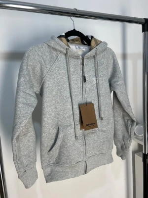  Burberry hoodie  - Snygg gråmelerad hoodie från Burberry med dragkedja, huva med dragsko och klassiska Burberry-detaljer i fodret. Mjuk bomullsblandning, två fickor fram och diskret logga på bröstet. Perfekt för en chill och stilren look.