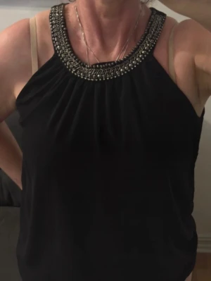 Svart topp med pärldetaljer från MyWear - Snygg svart ärmlös topp  med pärlbroderad halsringning som ger en lyxig känsla. Toppen har en lös passform och är tillverkad i ett lätt, mjukt material som faller fint. Perfekt för dig som vill ha något extra till outfiten.