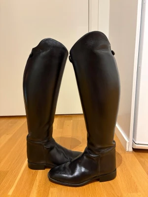 Cavallo Ridstövlar Grand Prix - Säljer ett par klassiska svarta ridstövlar i äkta läder. De är höga med rundad tå och har en stilren design utan dragkedja. Insidan är ljusare och stövlarna har en knappdetalj upptill. Perfekta för dig som vill ha en clean look till ridningen.