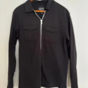 Svart overshirt med dragkedja MAN - Stilren svart overshirt från MAN med dragkedja framtill och två bröstfickor med lock. Overshirten har klassisk krage och raka ärmar, perfekt för lager på lager. Materialet känns mjukt och bekvämt, passar till många olika looks. Längd: 70cm, P2P: 48cm
