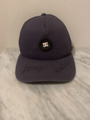 Svart DC keps med autografer från Kebbe och issa - Säljer en svart keps från DC med rund logotyp framtill. Kepsen har böjd skärm och autografer från keebabb och Isabelle på framsidan. Insidan av skärmen har ett holografiskt klistermärke med texten 'Jag är i min prime'. Klassisk och stilren modell i tyg.