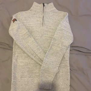 Ljusgrå stickad tröja Superdry - Ljusgrå stickad tröja från Superdry med halv dragkedja och hög krage. Tröjan har ribbade muddar vid ärmslut och nederkant samt en diskret Superdry-patch på ena ärmen. Tillverkad i mjukt stickat material, perfekt för kyliga dagar. 