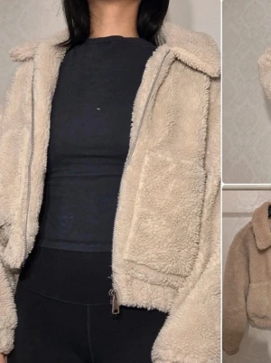 Beige teddyjacka från Urban Outfitters - Mysig beige teddyjacka med dragkedja framtill, stor krage och två stora fickor. Jackan har en boxy passform och är gjord i fluffigt teddy-material som ger en trendig och avslappnad look. Perfekt för att hålla värmen under kyliga dagar.