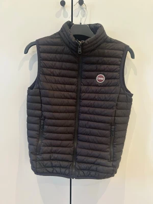 Svart dunväst från Colmar - Snygg svart dunväst från Colmar med quiltad design och hög krage. Västen har två dragkedjefickor framtill och Colmar-logga på bröstet. Perfekt att slänga över en hoodie eller tröja för extra värme. Passar ungefär 160-170 cm lång. Skriv om du är intresserad.