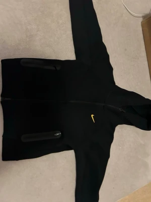 Svart Nike NOCTA set med huva - Snyggt svart set från Nike NOCTA med gul logga. Setet består av en hoodie med dragkedja och två fickor samt matchande byxor med dragsko och NOCTA-detaljer. Båda plaggen har diskreta svarta detaljer och är tillverkade i mjukt material. 500 för tröjan och byxorna 