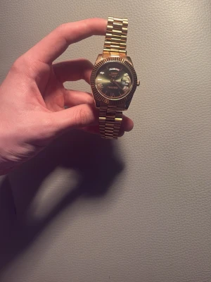 Guldfärgad klocka med länkarmband - Stilren guldfärgad klocka med klassiskt länkarmband i metall. Urtavlan är mörk grön med romerska siffror och dag- och datumvisning. Klockan har en markerad räfflad kant runt urtavlan som ger en lyxig känsla.