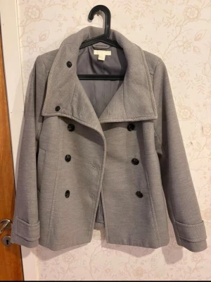 Ljusgrå skepparkavaj från H&M - Stilren ljusgrå skepparkavaj från H&M med dubbelknäppning och stora svarta knappar. Klassisk krage och två sidofickor. Tillverkad i ett mjukt tyg som ger en snygg och modern look. Perfekt för dig som vill ha en tidlös jacka med trendig känsla.