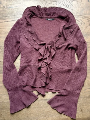 Plommonlila volangkofta från Gina Tricot - Säljer en plommonlila/burgandy kofta från Gina Tricot med volangdetaljer längs hela öppningen och vida trumpetärmar. Koftan är perfekt för dig som gillar romantiska detaljer - även perfekt för alla❤️dag.