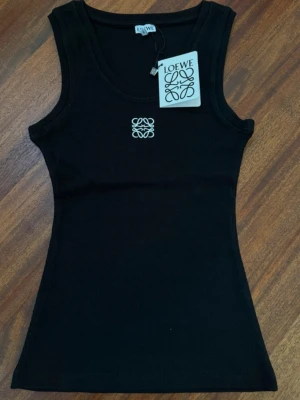 Svart ribbad linnetopp från Loewe - Snygg svart ribbad linnetopp från Loewe med broderad vit logga framtill. Toppen är ärmlös och har en tight passform, perfekt för att styla med jeans eller kjol. Materialet känns mjukt och stretchigt, vilket gör den superbekväm.