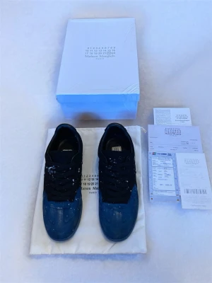 Maison Margiela blå-svarta sneakers - Unika sneakers från Maison Margiela i svart och blått med vit färgstänk-design. Lågt skaft, svarta skosnören och ovandel i en mix av mocka och läder. Snygg kontrast mellan färgerna och den konstnärliga finishen gör dem till ett statement-piece för dig som gillar streetwear.