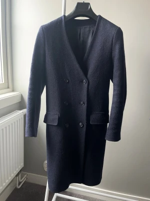 Mörkblå ullkappa med dubbelknäppning från Filippa K, st. XS - Stilren mörkblå kappa i ull med dubbelknäppning framtill och två fickor med lock. Kappan har en rak passform, V-ringad hals och ett bälte baktill för extra detalj. Perfekt för kyliga dagar och ger en klassisk look. Lagad i foder (se sista bild) men inget som syns på eller påverkar funktionen. Nypris 4000:-