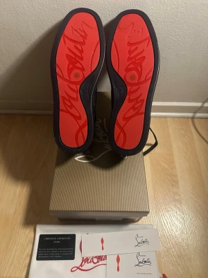 Christian Louboutin sneakers navy - Snygga sneakers från Christian Louboutin i mörkblå mocka med ikoniska röda sulor och nitar på sidorna. Skorna har en låg profil och klassisk siluett med exklusiva detaljer och logotyp på sulan. Perfekta för dig som vill sticka ut med stil. Svart mörkblå har även svarta