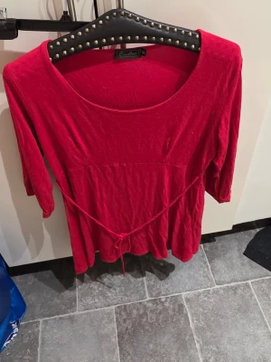 Röd topp med knyt från Ecoline - Snygg röd topp från Ecoline med rund halsringning och knytband i midjan. Modellen har trekvartsärmar och är tillverkad i ett mjukt material som känns skönt mot huden. Perfekt för dig som gillar färg och vill ha en enkel men stilren look.