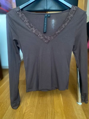 Brun långärmad topp med spets - Mörkbrun långärmad topp från The Black Label Collection med v-ringning dekorerad med spetsdetaljer. Tillverkad i mjukt material som sitter bekvämt och har en stilren, enkel design som passar till många olika looks.Använd en gång