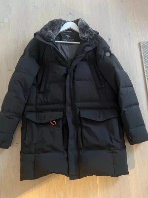 Svart parkas från Snoot med fuskpäls - Stilren svart parkas från Snoot med mjuk fuskpäls i kragen och stora fickor framtill. Jackan har vadderad design, dragkedja och tryckknappar, samt en diskret logotyp på ärmen. Perfekt för kalla dagar och riktigt snygg till vintern. Modellen är Agrigento Black. Väldigt varm och i princip oanvänd