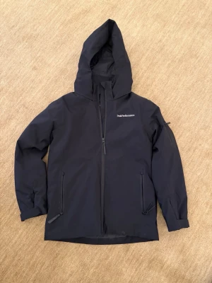 Svart skidjacka från Peak Performance Maroon - Snygg svart vindjacka från Peak Performance med huva och dragkedja framtill. Jackan har två snedställda fickor med dragkedja och diskret logga på bröstet. Tillverkad i ett lätt och tåligt material som står emot vind.