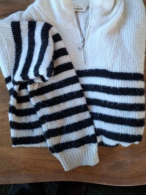 Randig stickad tröja från KappAhl - Stickad tröja från KappAhl i vitt och svart med breda horisontella ränder. Tröjan har halv dragkedja framtill och hög krage. Materialet ser mjukt och mysigt ut, perfekt för kyligare dagar. Långa ärmar med ribbade muddar.