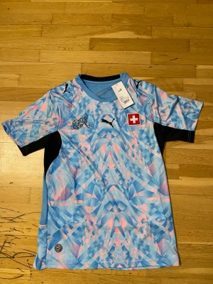 Schweiz fotbollströja Puma blå - Snygg Schweiz fotbollströja från Puma i blått med rosa och vita mönster. Tröjan har korta ärmar, rund hals och Schweiz flagga på bröstet. Tillverkad i återvunnet material (RE:FIBRE). Perfekt för fotbollsträning eller match! Priset kan diskuteras 🤝