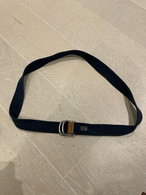 Mörkblått bälte från Polo Ralph Lauren - Snyggt mörkblått bälte från Polo Ralph Lauren med dubbla silverfärgade D-ringar och en brun läderdetalj vid änden. Bältet är gjort i ett slitstarkt tygmaterial och har en klassisk, stilren look med Polo-logga på etiketten. Org pris: 800kr