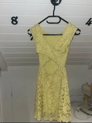 Gul spetsklänning med cut-out - Söt gul kortklänning i spets med blommigt mönster och snygg cut-out detalj framtill. Klänningen har ärmlös design och rundad halsringning. Perfekt för sommarens alla tillfällen!