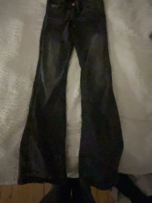 Svarta bootcut jeans med utsvängda ben - Säljer ett par svarta bootcut jeans med utsvängda ben och klassisk femficksdesign. Jeansen har låg midja och subtila slitningar framtill. Perfekta för dig som gillar en retro vibe och vill ha ett par jeans som sticker ut lite extra.