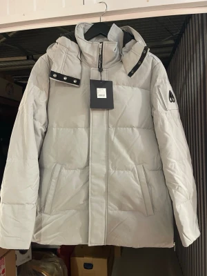 Ljusgrå dunjacka från Moose Knuckles - Snygg ljusgrå dunjacka från Moose Knuckles med avtagbar huva och metallogo på ärmen. Jackan har hög krage, dragkedja framtill och två snedställda fickor. Perfekt för kalla vinterdagar och riktigt stilren look.