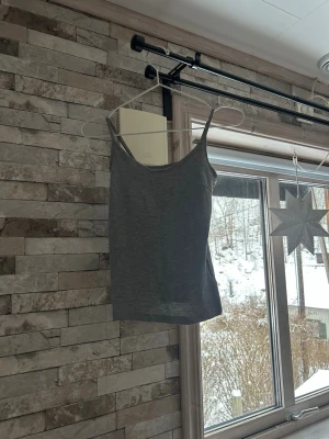 Grå linne med smala axelband - En enkel och stilren grå linne med smala axelband och rundad halsringning. Perfekt att matcha med jeans eller kjol för en avslappnad look. Mjuk och bekväm, passar till många olika outfits.