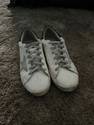 Vita sneakers med stjärndetalj - Säljer ett par vita Golden Goose sneakers med grå stjärna på sidan och svart hälparti. Skorna har klassisk låg profil, vita snören och slitna detaljer för en cool, avslappnad look. Tillverkade i läder och mocka för en lyxig känsla.