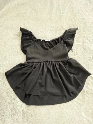 Svart volangblus med puffärm - Snygg svart blus med volang nertill och puffiga korta ärmar. Blusen har en lätt v-ringad halsringning och är tillverkad i ett mjukt tyg som ger ett fint fall. Perfekt för dig som gillar en feminin och trendig stil.