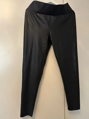 Svarta leggings i fuskskinn - Snygga svarta leggings i fuskskinn med hög midja och stretchigt material. De har en slät yta och smal passform som ger en cool look. Perfekta att styla med oversized tröja eller hoodie.