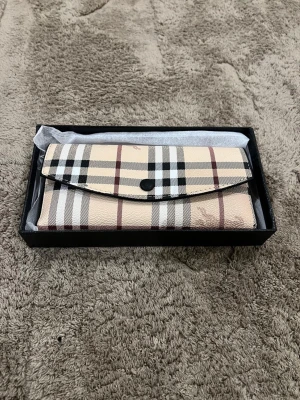  plånbok i klassiskt rutmönster - Snygg plånbok i beige med det ikoniska rutmönstret och svarta detaljer. Plånboken är tillverkad i läder och har flera kortfack på insidan. Stängs med en knapp framtill och har guldtext med logga inuti.