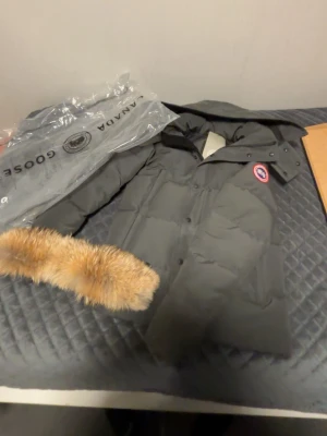Svart dunjacka från Canada Goose - Säljer en svart dunjacka från Canada Goose med avtagbar pälskant på huvan och klassisk logotyp på ärmen. Jackan har dragkedja och knappar framtill samt en stilren, rak passform. Perfekt för kalla vinterdagar.