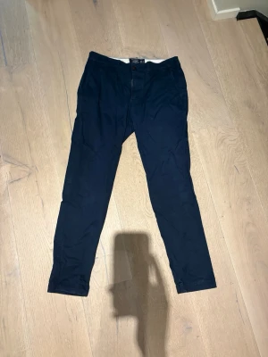 Mörkblå slim chinos Abercrombie - Snygga mörkblå chinos från Abercrombie & Fitch i modellen Langdon Slim. Byxorna har en smal passform, klassisk knappstängning och är tillverkade i ett mjukt bomullsmaterial. Perfekta för en clean och stilren look.