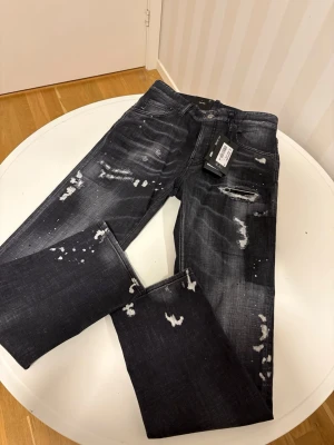 Svarta distressed jeans från DSQUARED2 Slim Fit - Snygga svarta jeans från DSQUARED2 med slitna detaljer, färgstänk och trendig tvätt. Modellen har klassisk femficksdesign och knappgylf. Perfekta för dig som gillar en edgy streetstil och vill sticka ut med dina jeans. Byxorna är helt nya, har aldrig använts fick dom i present idag och dom va för stora. Dom är 100% kan visa bevis till seriösa personer. Priset är 2999kr dom kostar nästan 7000kr i butiken, 