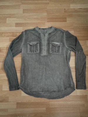 Grå henley med bröstfickor - Snygg grå henley-skjorta med lång ärm och två bröstfickor med lock. Skjortan har knappar vid halsen och en avslappnad passform. Perfekt för en chill och stilren look. Bomull 70%, silk 30% Bröst 104cm, kan passa även M