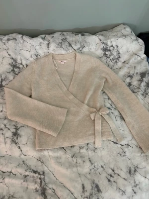Beige omlottstickad tröja med knyt - Mysig beige stickad omlott-tröja med knytband i midjan och långa ärmar. Tröjan har ribbstickade muddar och en djup v-ringning framtill. Perfekt för lager-på-lager och en chill look.