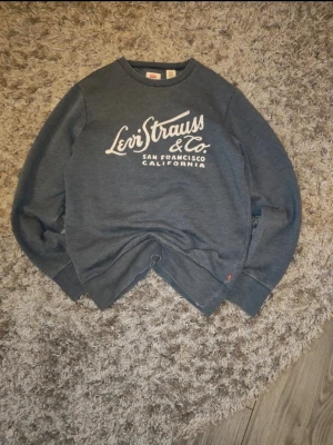 Blå sweatshirt från Levi's med tryck, storlek S - Säljer en blå sweatshirt från Levi's med stort vitt tryck på bröstet. Tröjan har rund hals, långa ärmar och är gjord i mjuk bomullsmix. Inget tryck på ryggen. Använd men i fint skick.