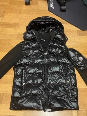 Svart dunväst med huva från Moncler - ‼️‼️pris kan diskuteras Snygg svart dunväst från Moncler i glansigt material med huva och dragkedja framtill. Västen har två fickor med tryckknappar och en Moncler-logga på ärmen. Perfekt för kalla dagar när du vill ha både stil och värme.